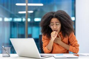 A woman coughs while working at a desk, wearing glasses and an orange shirt, in a modern office setting. photo
