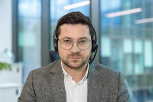 Close-up portrait of a young serious male office worker and assistant sitting at his workplace in a headset and looking intently at the camera. photo