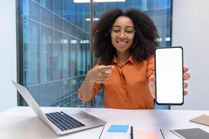 A smiling woman with afro hair points at a blank smartphone screen in an office setting, near a laptop. photo