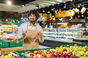 A smiling grocery store worker holds apples, standing in front of a display of fresh produce at a supermarket. photo