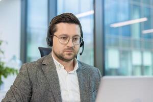 Close-up photo of a young man in a suit and headset working seriously in the office on a laptop.