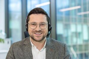 Close-up portrait of a young male office worker sitting at his workplace in a headset and suit and looking at the camera with a smile. photo