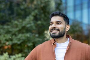 A smiling Indian man with a beard, dressed in a casual orange shirt, looks off-camera with green foliage in the background. photo