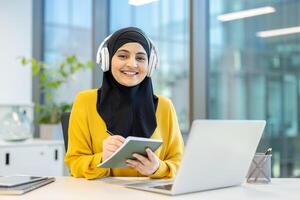 A smiling Muslim woman wearing a hijab and headphones, working on a laptop and taking notes at her desk in a modern office. photo
