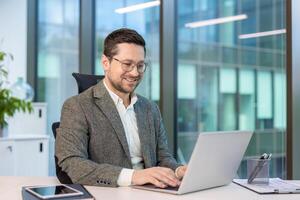 Smiling young businessman sitting in a suit in the office at a table and working on a laptop. photo