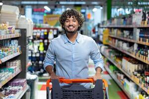 A smiling man pushes a shopping cart down an aisle in a brightly lit grocery store, focused on purchasing. photo
