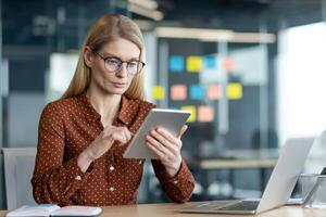 A businesswoman is working at her desk with a tablet and laptop in a modern office setting, wearing glasses and a button-down shirt. photo