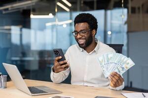 A smiling Black man in glasses holding money and looking at his phone, likely at good news in an office setting. photo