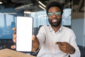 An African American man in glasses points to a blank smartphone screen, smiling confidently at the camera in an office setting. photo