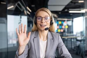 A businesswoman in a suit and headset waves during a call, smiling brightly. The office background suggests a collaborative work environment. photo