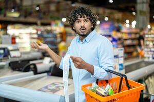 A shocked man looks at his grocery bill, holding a shopping basket in a supermarket. He is surprised by the total. photo