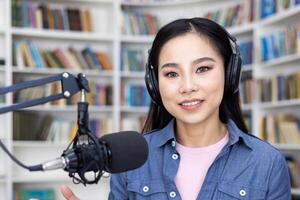 A young woman with headphones on speaks into a microphone in front of bookshelves, recording a podcast. photo