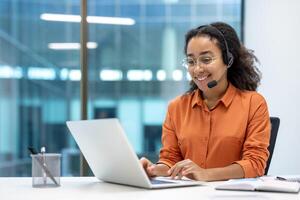 Smiling customer service representative wearing a headset and glasses working on a laptop in a modern office setting. Focus on communication and support. photo