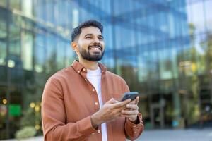 A smiling man holds a smartphone in front of a modern glass building, suggesting a business or technology context. photo