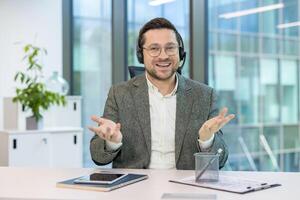 Portrait of a young smiling man sitting at a desk in an office, wearing a suit and headset, explaining and talking to the camera. photo