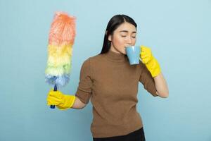 Asian housekeeper holding colorful duster and drinks coffee while taking a break during spring cleaning. Female cleaner standing against blue background enjoying moment of rest from chores. photo