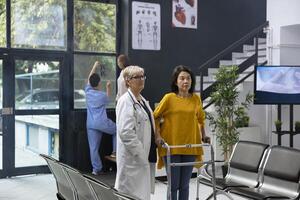 Doctor assisting patient while she uses walking frame during rehabilitation therapy, having a checkup appointment. Physical therapy discussion with mobility issues, healing process. photo