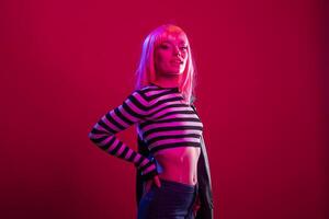 Female model with striped outfit and pink wig stands in calm yet assertive pose against isolated background, exuding vibrant energy. Glamorous lady with hand on waist, radiating confidence in studio. photo