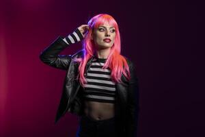 Young fashionable female with pink bangs hairstyle and eyeliner stands confidently with edgy outfit in studio portrait. Glamorous caucasian lady in leather jacket and crop top, posing with raised arm. photo