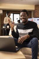Smiling male manager sits on couch, greeting remote coworkers with a wave during online call. Businessman participates in virtual meeting from cozy office, connecting with company stakeholders. photo