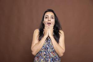 Stunned Indian woman with hands on cheeks and eyes wide open, expressing dramatic disbelief. Beautiful female model stands against brown background, looking at camera in shock and amazement. photo