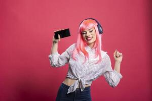 Excited woman with pink hair and headphones, enjoying music through cellphone. Cheerful young lady in casual outfit, dancing and having great time in studio, standing against isolated background. photo
