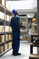 Inventory system managed by african american worker in safety gear, checking the cargo boxes on the racks. Storage units hold packages and containers for delivery, import export. photo