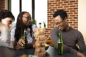 African american couple drinking beer and making goofy facial expressions in brick wall room. Group of friends at table, enjoying board games and relaxed bonding time with wooden game pieces. photo