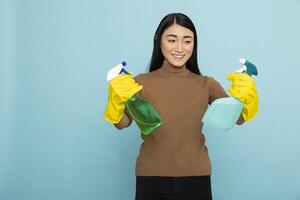 Smiling asian janitor standing against blue background, confidently holding spray bottles and wearing yellow rubber gloves. Happy housekeeper ready to begin morning housecleaning routine. photo