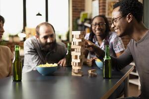 Young black man with glasses, happily pulling wooden block from jenga tower during indoor game session. Diverse group of friends watch intently, enjoying shared moments in brick wall room. photo