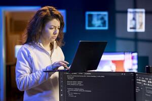 IT system administrator doing software debugging using laptop, checking glitches in lines of code displayed on desktop PC monitors. Woman standing in office at home fixing coding issues using notebook photo