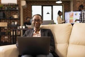 Smiling black female entrepreneur works from cozy office lounge, using laptop to analyze business data. Professional woman checking emails and preparing for productive day with her personal computer. photo