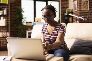 Focused woman works from home, reviewing company growth analytics on laptop. African American female entrepreneur touching device screen and reading online data with full concentration. photo
