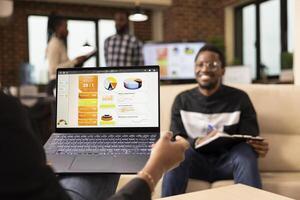African american entrepreneur holding laptop showing company analytics, reviewing marketing strategy with male coworker. Selective focus on device held by businessperson, showing performance graphs. photo
