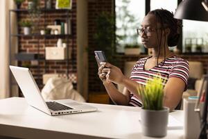 Casually dressed woman multitasks from home, checking messages on smartphone while working on laptop. Focused black freelancer with glasses, sitting at desk and using mobile device. photo