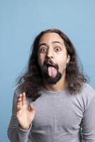 Goofy young adult with long hair and beard poses playfully in the studio, expressing positive emotions and carefree humor. Joyful person showcases unique, comical personality and fun spirit. photo