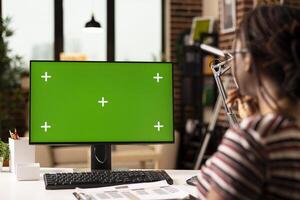 Freelancer works from home using computer with green screen, reviewing template for editing or presentation. Female remote employee sits at desk with clipboard, looking at isolated chroma key display. photo