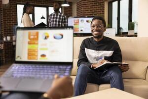 Two business professionals meet in office to share ideas and collaborate on project task. African american sales manager with laptop presents commercial offer to startup company client seated on sofa. photo