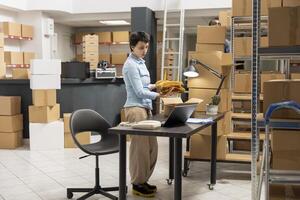 Woman depot worker preparing boxes for shipment in local brand supply room, managing the orders on a laptop. Fulfillment center focused on retail operations and e commerce distribution. photo