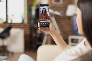 Remote employee sits on sofa, looking at digital device screen during online collaboration with white businesswoman. Female manager discussing marketing strategy with team member on call. photo