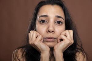 Closeup of sad indian woman looking directly at camera with troubled, distressed expression. Anxious female model resting head on her hands, feeling emotional after receiving upsetting news photo