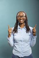 Smiling african american woman points upward with both index fingers, highlighting space for promotional content. Black female model in blue shirt, looking at camera and gesturing above. photo
