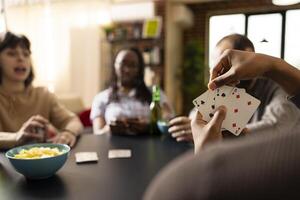 African american hand displaying winning card combo while others focus on game. Young adults seated at table with beer bottles and snacks, having relaxed bonding time, playing games. photo