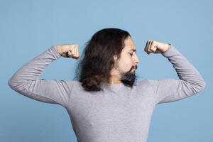 Athletic young guy flexing his arms muscles against blue background, showing off his toned fit physique in front of the camera. Fitness motivation and determination from a strong man. photo