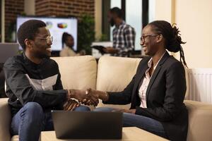 Happy businessman and businesswoman shaking hands near laptop while analyzing performance metrics. African american colleagues seated on couch, showcasing teamwork and excitement in marketing agency. photo