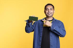 Confident young adult pointing at the green screen display on smartphone, pointing at the screen on the gadget. Indian person with positive mood against orange background in studio. photo