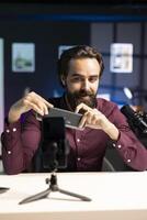 Cheerful online star doing technology review of wireless portable speaker, filming with phone on tripod. Happy influencer showcasing music playing device in apartment studio photo