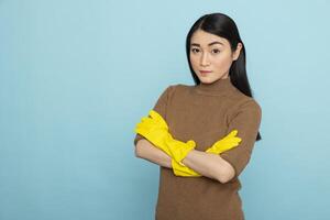 Portrait of female janitor wearing yellow gloves, standing confidently with crossed arms, ready to provide cleaning services. Asian housekeeper posing against blue studio background. photo