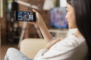 Businesswoman using cellphone, joins virtual shareholder meeting from apartment. Remote female manager sitting on sofa, listens and speaks with colleagues through mobile device in conference. photo