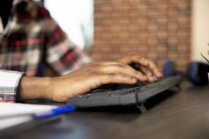 Closeup of african american individual using keyboard and updating company financial documents on desktop pc. Black man seated at home office desk, typing data in computer, preparing business project. photo